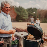 Man holding a drink standing next to a  grill with Rotisserie on it, outdoors.
