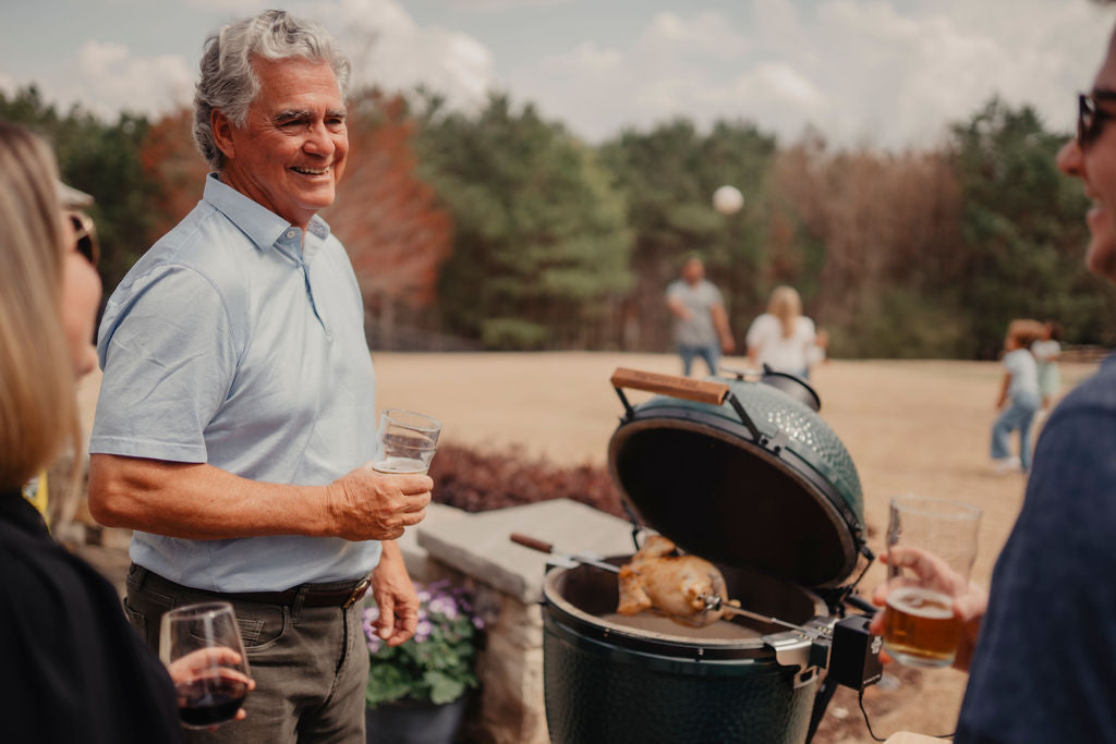 Man holding a drink standing next to a  grill with Rotisserie on it, outdoors.